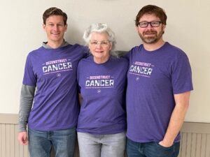 A mother and her two adult sons pose for a photo all wearing matching purple t-shirts.